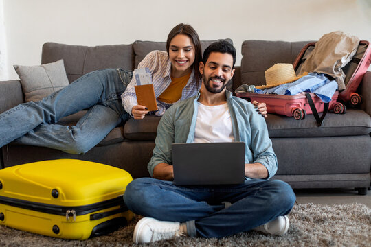 A young couple is sitting on the floor in a cozy living room, surrounded by luggage. They are looking at a laptop and appear to be planning their upcoming trip together. - Powered by Adobe