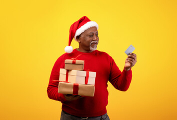 Merry senior black man in a Santa hat holds wrapped Christmas gifts and a credit card while shopping. The cheerful atmosphere shows his joy during the holiday season on an orange background.