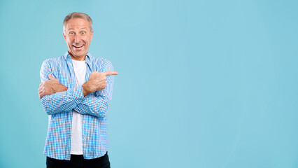 Mature man smiles widely while pointing at the side, promoting an empty space for ads. He stands in...