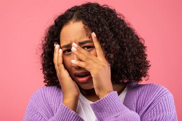 In a pink studio setting, a young black woman is visibly scared and covering her face in panic. She...
