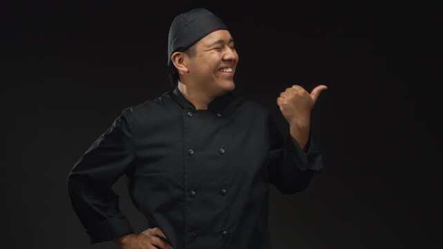 Man chef in black cook uniform pointing thumb to side with hand on hip in studio under black lighting; pride skill.