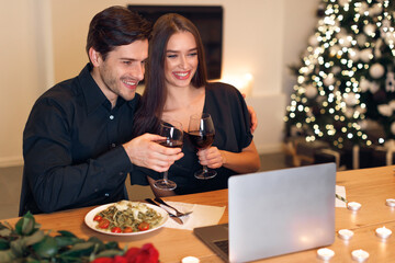Two people sit closely at a table, celebrating with wine and a delicious meal. They connect with friends via video call, surrounded by holiday decorations.