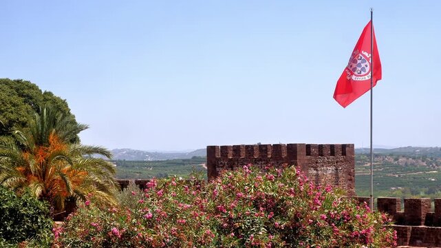 The Flag of Silves flies high above Silves Castle in the Algarve region of Portugal with palm tree and pink flowers