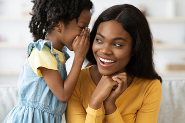 A young girl leans in to whisper to her smiling mother in a warm, inviting home setting. Their connection is filled with joy and playful energy, highlighting their close bond.