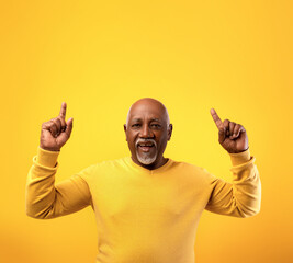 In a vibrant orange studio, an elderly African American man cheerfully points upwards with both...