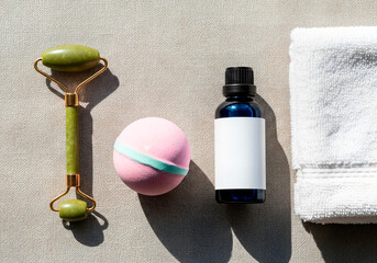 A flat lay of skincare products including a jade roller, a pink bath bomb, a blue essential oil bottle, and a white towel on a textured surface.