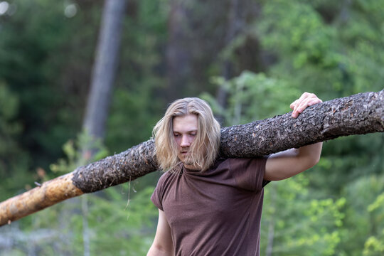 Exhausted, tired young man carrying tree trunk, log on his shoulder in a forest setting.