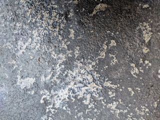 A dark, wet, granular surface, likely weathered asphalt or concrete, featuring a central area of lighter, flaking material. The image exhibits high contrast between the damp, deep gray background.
