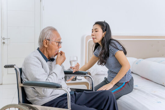 Young Caregiver Giving Pills and Water to Elderly Man in Wheelchair at Home