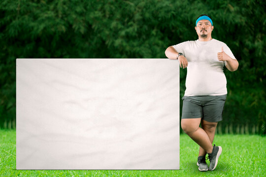 Portrait of an overweight man in exercise wear giving a thumbs up and leaning on a blank white billboard or sign in a park.