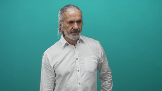 Man with long gray hair and beard wearing white shirt looking down in studio against teal background; pensive solitude.