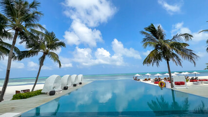 Infinity pool at a tropical resort with white sandy beach, turquoise ocean, and palm trees under a blue, cloudy sky