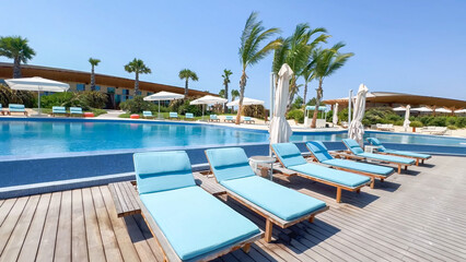 Resort swimming pool deck with wooden flooring and a row of lounge chairs with bright blue cushions under a clear sky