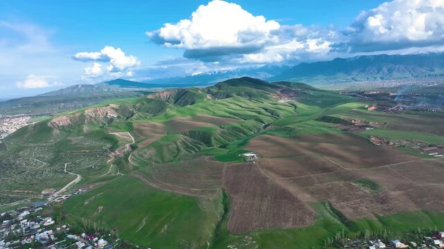 High aerial view over vast, contoured foothills with patches of green grass and cultivated brown fields, backed by snow-capped mountains in the distance