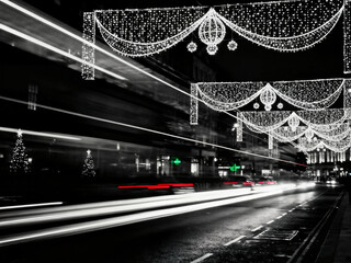 A dramatic black and white night long exposure of a city street during the Christmas season. Decorative lights hang over the road with streaks of light from moving traffic below.
