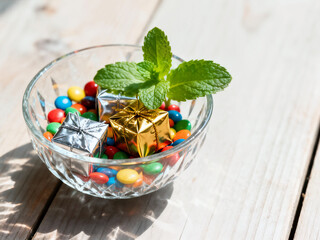 A small crystal bowl holds colorful candies and tiny silver and gold foil-wrapped gift boxes. A sprig of fresh mint leaves adds a touch of green on a wooden background.