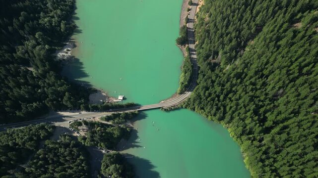 Top-down aerial drone shot showing a bridge and mountain road crossing the turquoise glacier water of Diablo Lake in North Cascades National Park. Kayaks float below on the vivid emerald water.