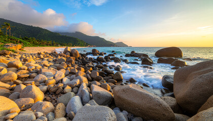 Golden hour light illuminates a rocky beach with waves crashing. Lush green hills meet the tropical shoreline under a warm, vibrant sky.