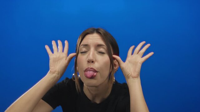 Young hispanic woman sticks tongue out and spreads hands in blue studio with silly expression; mischief playfulness.