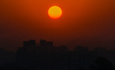 Fototapeta premium Beautiful sunset scene with a glowing orange sun above city buildings, creating a dramatic silhouette against the warm evening sky. The vibrant gradient tones of orange and red symbolize peace, hope