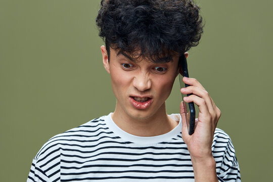 frustrated young man talking phone, curly hair, striped shirt, isolated green background, emotion of anger and confusion, casual style - Powered by Adobe