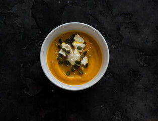 Pumpkin soup with cream cheese, pumpkin seeds in a white bowl on a dark background, top view