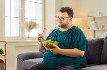 Fat overweight man eating salad with fork. In a bright living room, the male reflects on healthy...