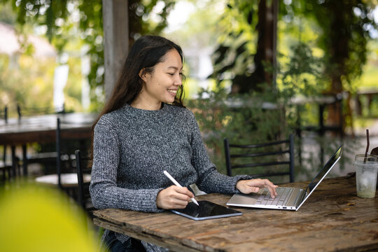 Asian freelancer smiling, multitasking with laptop and tablet at cafe
