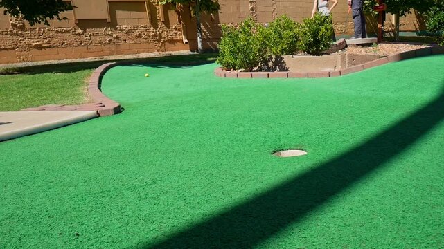 Couple playing mini golf and hitting a yellow golf ball around a curved corner towards the hole