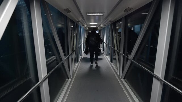 Slow motion follow shot of unidentified male passengers walking through jet bridge from airplane to airport terminal after early morning landing.