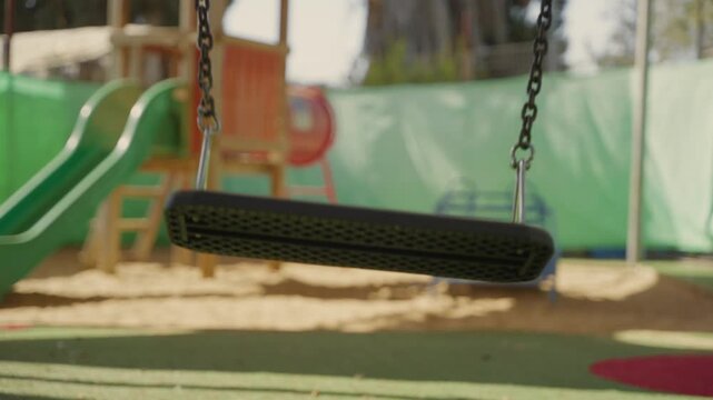 Empty swing moves gently in sunny playground with no children around. School or Kindergarten closure, neglect, missing child, bullying aftermath, or about safety concerns , supervision, child welfare.