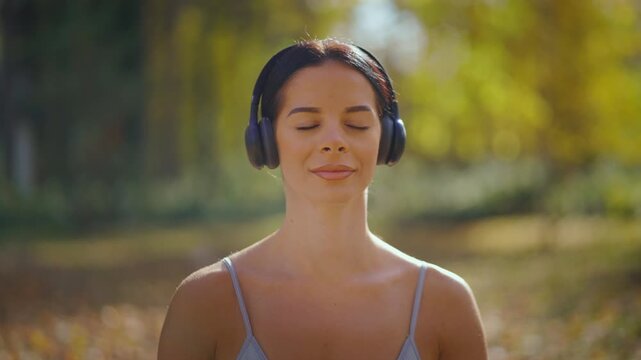 Serene woman with closed eyes wearing wireless headphones while meditating and practicing mindfulness, finding calm and relaxation in a beautiful, sunny park with a blurred background
