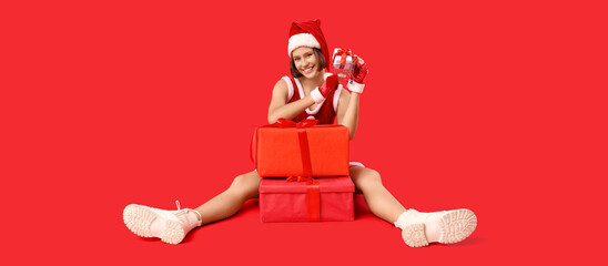 Beautiful young happy woman in Santa costume with shopping cart and gift boxes sitting on red background. Christmas celebration