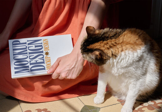 Woman in an orange dress holds a blank notebook while a cat curiously inspects the scene in warm sunlight. PSD template mockup