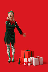 Happy young woman in elf costume with Christmas gift boxes pointing at something on red background