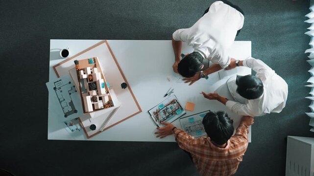 Group of skilled designer looking at model while discussing about building. Top view of architect engineer explain idea while talking and writing blue print scattering at meeting table. Alimentation.
