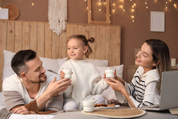 Happy family with glasses of milk and cookies lying on bed in winter evening at home