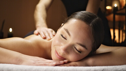 Obraz premium A woman receives a relaxing massage, eyes closed in serene contentment. The dimly lit setting with candles enhances the tranquil atmosphere.