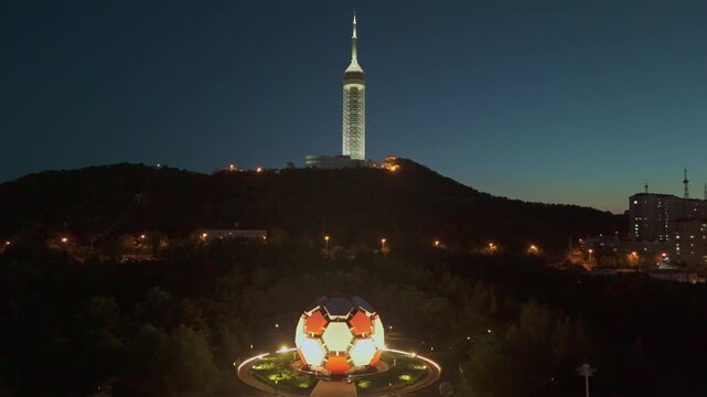 Orbit shot circling Laotong Park football building at sunset, showing surrounding city lights and urban layout