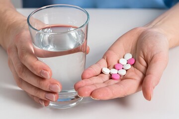 A glass of water in one hand and pills in the palm of the other hand, focused shot, bright lighting, blurred background.