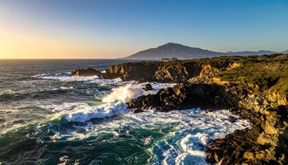 Crashing Waves On Rocky Coastline Under A Golden Sunset Sky With Distant Mountain Silhouette