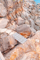 Wooden Walkway Amidst the Rocks: The image captures a wooden walkway delicately placed amidst the rugged embrace of towering rocks. Coastal rocks in soft pastel tones