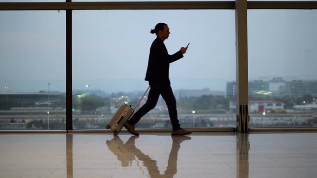 Man walking with trolley bag while using his smartphone inside airport terminal. Slow motion side view shot captures his silhouette against large window, with dusk sky and evening city visible outside