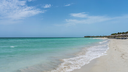 Fototapeta premium Beautiful tropical beach. The waves of the aquamarine ocean foam on the white sand. Rows of deck chairs under straw umbrellas. Blue sky, clouds. Cuba. Varadero. Resort. Hotel