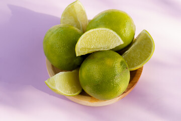 Fresh green limes and cut lime wedges arranged in a rustic wooden bowl on a light purple background with shadows