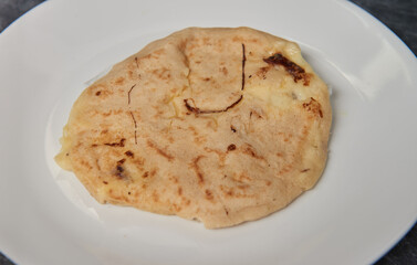 A close-up photo of a golden, cheese-filled pupusa on a white plate against a black-grey marble background.