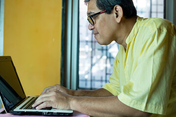 A man is sitting on the floor and works on a laptop at a desk near a window, concentrating on typing. The setting features bright yellow wall with natural light.