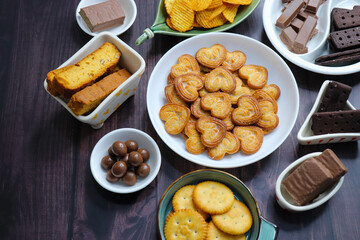 Assorted party snacks on table featuring chocolate cream sandwich biscuits, sugar coated heart cookies, wafer chocolates, chocolate coated caramel balls, salted crackers, and crispy potato chips.