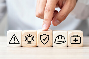 Naklejka premium Safety Management and Risk Protection Concept: Hand actively selecting shield with checkmark icon on wooden blocks surrounded by exclamation, alarm, hard hat and first aid kit, symbolizing proactive s