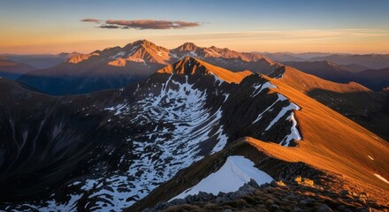 Aerial view of a rugged mountain range at sunset, casting warm light on snow-capped peaks